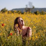 A woman sits with eyes closed, enjoying the sun in a vibrant yellow flower field.
