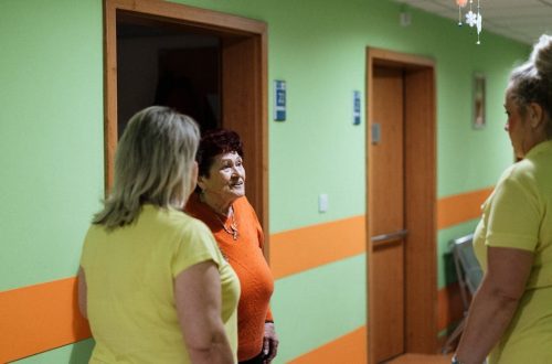 Senior woman engages in conversation with caregivers in a facility corridor.