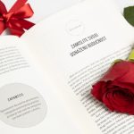 An open book with a red rose and a tied gift box next to it, conveying romance or celebration.